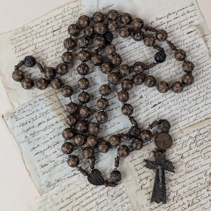 May include: A vintage rosary with dark brown beads and intricate detailing. The rosary features a wooden crucifix with a carved figure of Jesus. The beads are connected by a metal chain, and the rosary rests on aged paper with handwritten script.