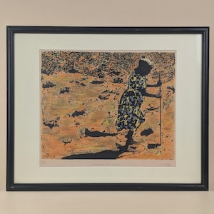 Può includere: Stampa incorniciata di una persona che cammina con un bastone, che indossa un vestito fantasia. L'opera d'arte presenta una tavolozza di colori neri, gialli e arancioni. La stampa è ambientata su uno sfondo beige e incorniciata in una cornice nera.