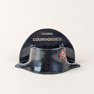 Puede incluir: Un soporte negro para botellas de coñac Courvoisier con forma de sombrero bicornio. El sombrero tiene la palabra "COGNAC" encima de "COURVOISIER" en letras doradas. Una roseta roja, blanca y dorada está en el lateral.
