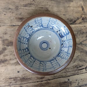 Può includere: Una ciotola rotonda in ceramica con un bordo marrone liscio e un interno bianco sporco maculato. L'interno presenta motivi blu dipinti a mano composti da linee verticali smerlate e punti, con un cerchio blu concentrico proprio al centro.