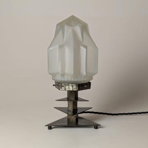 May include: An Art Deco table lamp with a frosted glass shade and a geometric base. The shade is a light, opaque white, and the base features triangular tiers. The lamp has a black cord. The lamp's height is approximately 25 cm.