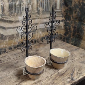 Op de afbeelding: Twee wandplantenhouders met zwarte smeedijzeren achterplaten met ingewikkelde krulpatronen. Elke houder bevat een cirkelvormige metalen ring die een beige, taps toelopende keramische pot met een gladde, geglazuurde binnenkant ondersteunt.