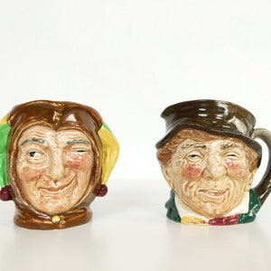 Puede incluir: Dos tazas de cerámica con caras extravagantes. Una taza presenta un bufón con un sombrero verde y amarillo y una cara marrón y blanca. La otra taza presenta un hombre con un sombrero marrón y una cara marrón y blanca.