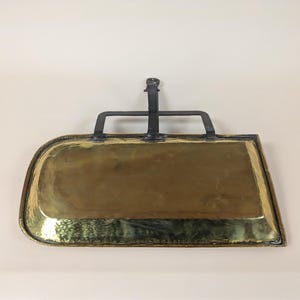 Pode incluir: Uma pá de lareira de latão com um cabo de metal preto. A pá tem uma forma retangular curva e é projetada para ser pendurada na parede.