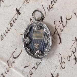 Puede incluir: Colgante antiguo de plata con marco ovalado y detalles ornamentados. El colgante presenta un fondo azul oscuro con un pequeño diseño floral de color claro y un trozo de tela tejida. El colgante está suspendido de un pequeño lazo.