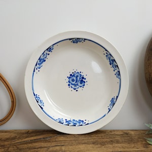 Peut inclure: Bol en céramique blanc avec un motif floral bleu. Le bol présente un groupe de fleurs central et une bordure d'accents floraux stylisés. Le bord est blanc et le bol est posé sur une surface en bois.