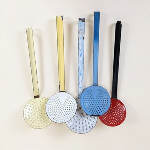 May include: Five vintage kitchen utensils with colorful enamel handles and perforated metal heads. The utensils are hanging from a white wall.