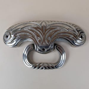 Puede incluir: Un tirador de cajón plateado con un diseño ornamentado de inspiración Art Déco. El tirador presenta un asa curva y una placa trasera decorativa con patrones florales y geométricos. El herraje es de metal y probablemente sea para un armario o cajón.