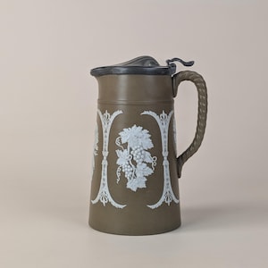 Puede incluir: Jarra verde oliva vintage con un diseño decorativo de uvas y hojas blancas. La jarra tiene un asa trenzada, una tapa gris oscuro y detalles decorativos blancos. Esta pieza antigua probablemente se usa para servir bebidas.