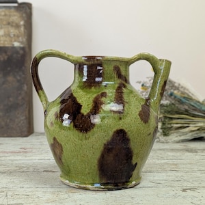 Puede incluir: Jarra de cerámica verde y marrón con dos asas y un pico. La jarra tiene un patrón moteado de manchas marrones oscuras sobre un fondo verde claro. La jarra está sobre una superficie de madera.