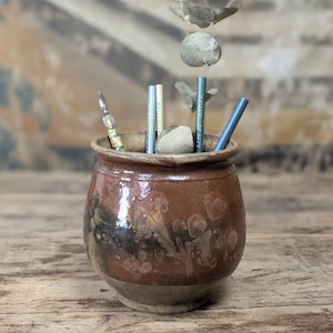 May include: A round, brown glazed ceramic pot with a wide rim, containing several pens and a small dried plant stem. The pot features a mottled, earthy finish with darker brown and tan patches, and the pens include one labeled Arts Decoratifs.
