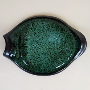 Può includere: Un piatto da portata in ceramica verde e nero con una superficie unica e testurizzata. Il piatto ha la forma di un pesce e ha una finitura lucida.