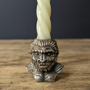 May include: A silver-colored candle holder with a detailed bust of a gorilla. The candle holder has a textured finish and holds a twisted, off-white candle. The candle holder is on a wooden surface.