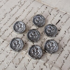Op de afbeelding: Een verzameling van zeven antieke zilverkleurige metalen knopen. Elke knoop heeft een gedetailleerd ontwerp van drie eikels en bladeren in een cirkelvormig frame. De knopen zijn gerangschikt op een vintage papieren achtergrond met handschrift.