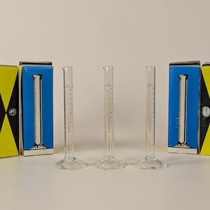 May include: Three clear glass graduated cylinders with a white scale and a hexagonal base. The cylinders are sitting on a white surface. The cylinders are packaged in blue and yellow boxes with the text "STABIL CAVALIER" and "GLASSEXPORT" printed on them.