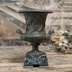May include: A dark bronze-toned metal urn featuring relief carvings of a winged figure and a seated person. The vessel has two handles shaped like animal heads, a wide fluted rim, and a square base made of mottled gray and black marble stone.