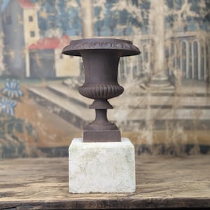 May include: A dark brown cast iron urn with a wide, circular rim featuring a decorative embossed pattern, a fluted bowl section, and a slender pedestal base. The urn is mounted on a solid, square, off-white stone block with a weathered texture.
