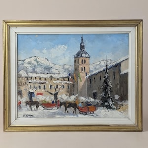 May include: Oil painting of a snowy village scene with a church tower, mountains, and horse-drawn sleighs. The artwork is framed in a gold-colored wooden frame. The painting has a winter theme with a blue sky and snow-covered buildings.