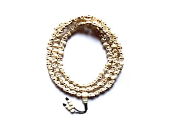 May include: A white skull bead mala necklace with a black tassel. The necklace is made of small, white skull beads that are strung together on a black cord. The necklace is approximately 18 inches long and has a black tassel at the end.