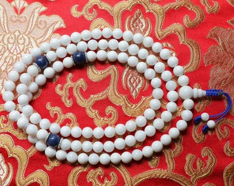 May include: White prayer beads with blue accents. The beads are strung on a cord with a tassel. The beads are arranged in a circle on a red and gold patterned fabric.