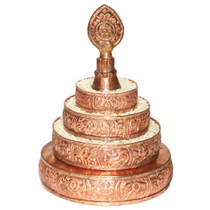 May include: A copper-colored, tiered, decorative object with intricate designs. The top tier features a pointed finial with a circular design. The object is likely a Tibetan prayer wheel.