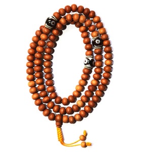 May include: A brown wooden mala bead necklace with three black and white eye beads. The necklace is strung with a yellow cord and has a tassel at the end.