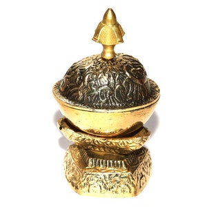 Puede incluir: Un soporte decorativo de metal dorado con una parte superior redonda y tres patas. La parte superior del soporte tiene un diseño adornado y un remate puntiagudo.