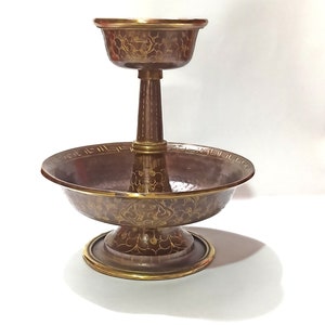 Puede incluir: Un plato de servir de latón de dos niveles con un acabado martillado y diseños florales intrincados en oro. El nivel superior es un tazón pequeño, y el nivel inferior es un tazón más grande con una base ancha.