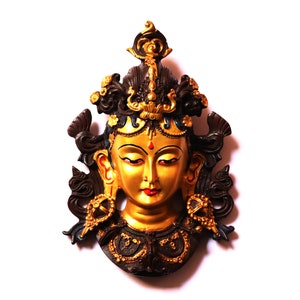 May include: A golden mask with a female face, intricate details, and a black and gold crown. The mask has a red dot on the forehead and red lips.