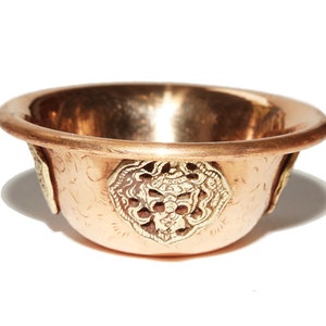 Puede incluir: Un pequeño cuenco de cobre con diseños intrincados y acentos dorados. El cuenco tiene una superficie lisa y pulida y está decorado con cuatro motivos florales ornamentados.
