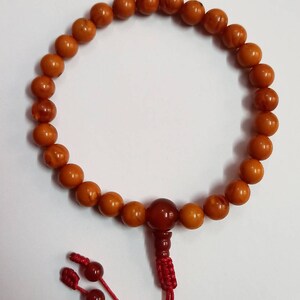 May include: A prayer bead bracelet featuring round, amber-colored beads. The bracelet has a larger, darker bead and a red tassel with two smaller beads. The beads are strung together, forming a circular shape.