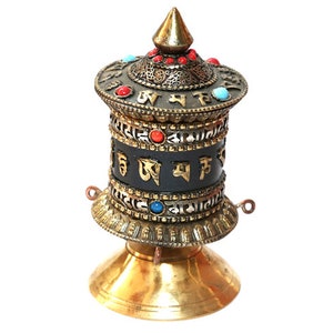 Könnte beinhalten: Ein goldenes Gebetsrad mit einem schwarzen Zylinder und bunten Edelsteinen. Das Rad ist mit komplizierten Mustern und tibetischer Schrift verziert.