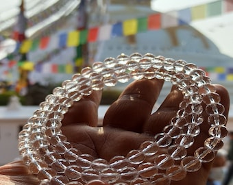 May include: A clear crystal mala with a white string tassel. The mala is made of round beads and is held in a hand. The background is a blurry image of a white building with colorful flags.
