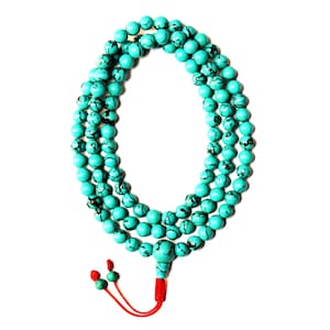 Puede incluir: Un collar de cuentas mala de color azul turquesa con un borla roja. Las cuentas son redondas y tienen marcas negras.