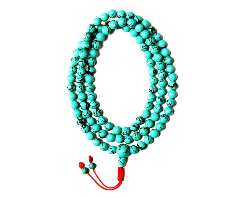 May include: A turquoise blue mala bead necklace with a red tassel. The beads are round and have black markings.
