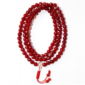 May include: A red gemstone mala with a clear crystal bead at the end and a red tassel. The mala is made of faceted beads and is strung on a red cord.