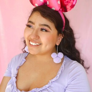 May include: A woman wearing a pink sequined headband with white polka dots and large pink bow ears. She is also wearing lavender earrings shaped like a mouse head. The woman is wearing a lavender top.