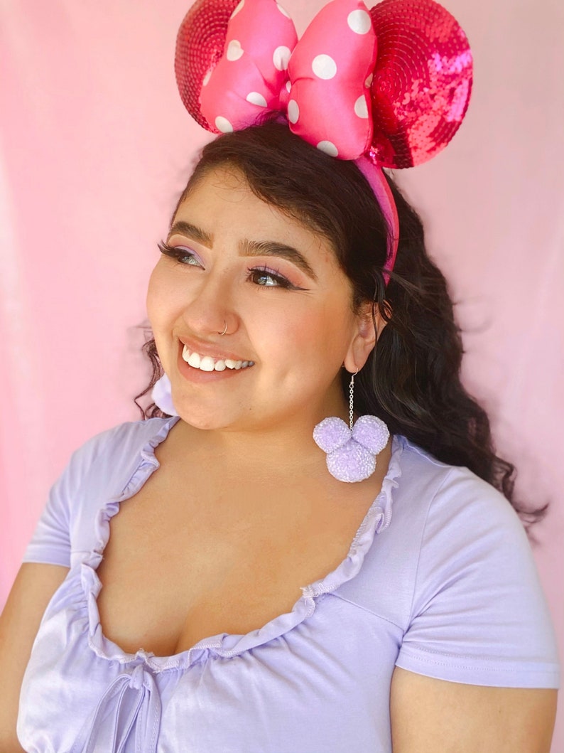 May include: A woman wearing a pink sequined headband with white polka dots and large pink bow ears. She is also wearing lavender earrings shaped like a mouse head. The woman is wearing a lavender top.
