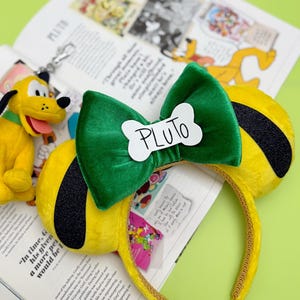 May include: Yellow and black Pluto-themed mouse ears with a green bow and a white bone-shaped tag that says "PLUTO". The headband is gold and rests on an open book with a Pluto plush toy.