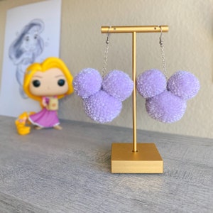 May include: A pair of lavender pom-pom earrings shaped like a mouse head. The earrings hang from silver-colored chains and hooks. The earrings are displayed on a gold-colored stand. A figurine of a cartoon character is in the background.