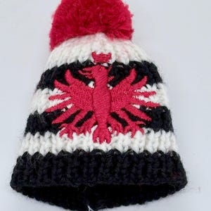 May include: A chunky knit beanie featuring horizontal stripes in black and white. The hat is topped with a large red pom-pom and displays a prominent embroidered red eagle emblem centered on the front, showcasing detailed stitching and texture.