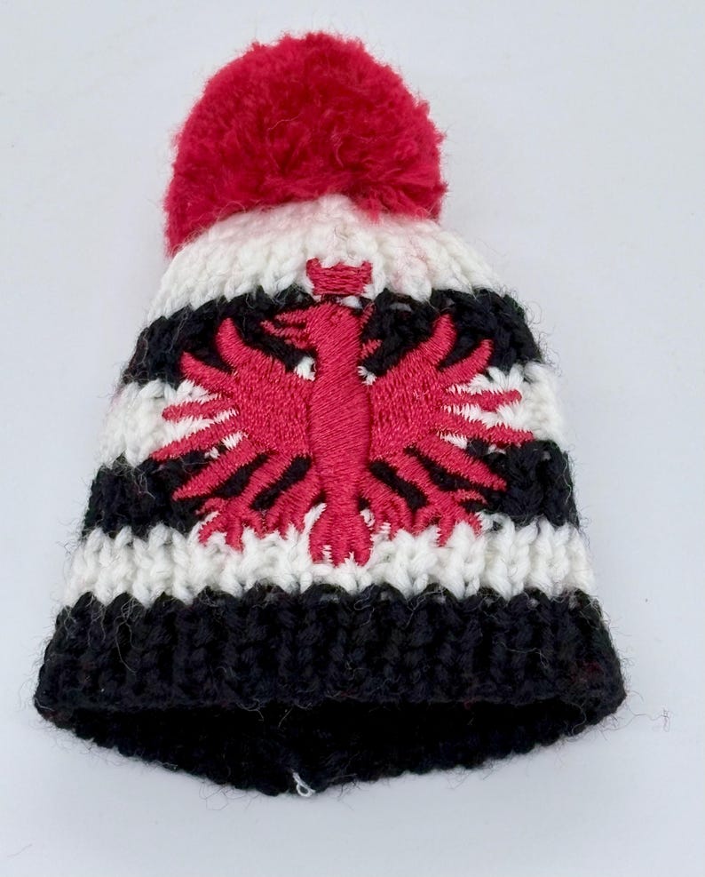May include: A chunky knit beanie featuring horizontal stripes in black and white. The hat is topped with a large red pom-pom and displays a prominent embroidered red eagle emblem centered on the front, showcasing detailed stitching and texture.