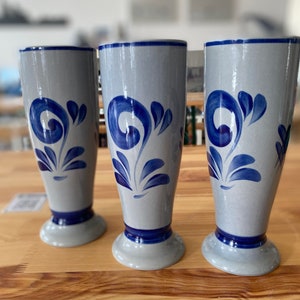 May include: Three grey ceramic beer steins with blue floral designs. The steins are tall and slender with a rounded base.