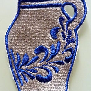 Puede incluir: Un parche bordado azul y blanco de una jarra de cerveza alemana tradicional con un dise&ntilde;o floral.