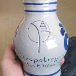 Könnte beinhalten: Weißer Keramikkrug mit blauen Akzenten und einem blauen Logo. Der Krug hat einen runden Körper und einen schmalen Hals, mit blauen Streifen oben und unten. Text ist auf dem Krug sichtbar.