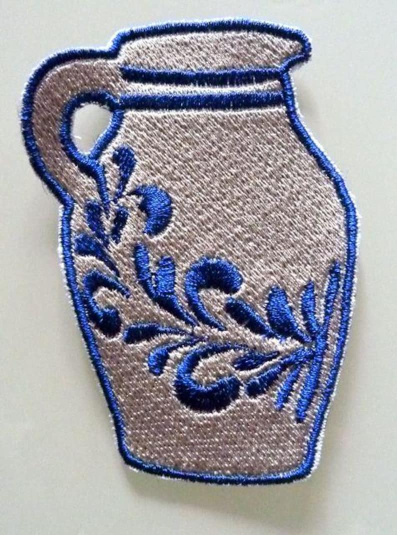 Puede incluir: Un parche de planchado bordado con un dise&ntilde;o tradicional de jarra de gres alemana, con un dise&ntilde;o floral azul y blanco.
