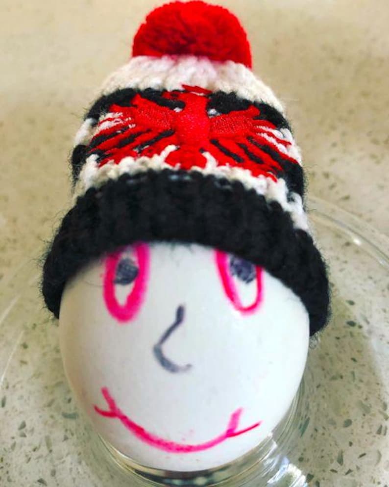 May include: A white egg decorated with a hand-drawn face featuring pink eyes, a gray nose, and a pink mouth, wearing a small, hand-knitted beanie. The beanie is white with a black brim, a red pom-pom, and a red double-headed eagle emblem.