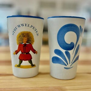 Op de afbeelding: Twee witte keramische mokken met blauwe rand. Een mok heeft een cartoonillustratie van een persoon met een rood shirt en de tekst "Struwwelpeter". De andere mok heeft een blauw bloemenmotief.