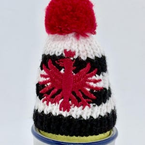 May include: A small knitted hat with a red pom-pom on top, featuring horizontal stripes of white and black yarn. The center of the hat displays a red embroidered eagle emblem. The hat is placed on top of a small, cylindrical ceramic container.