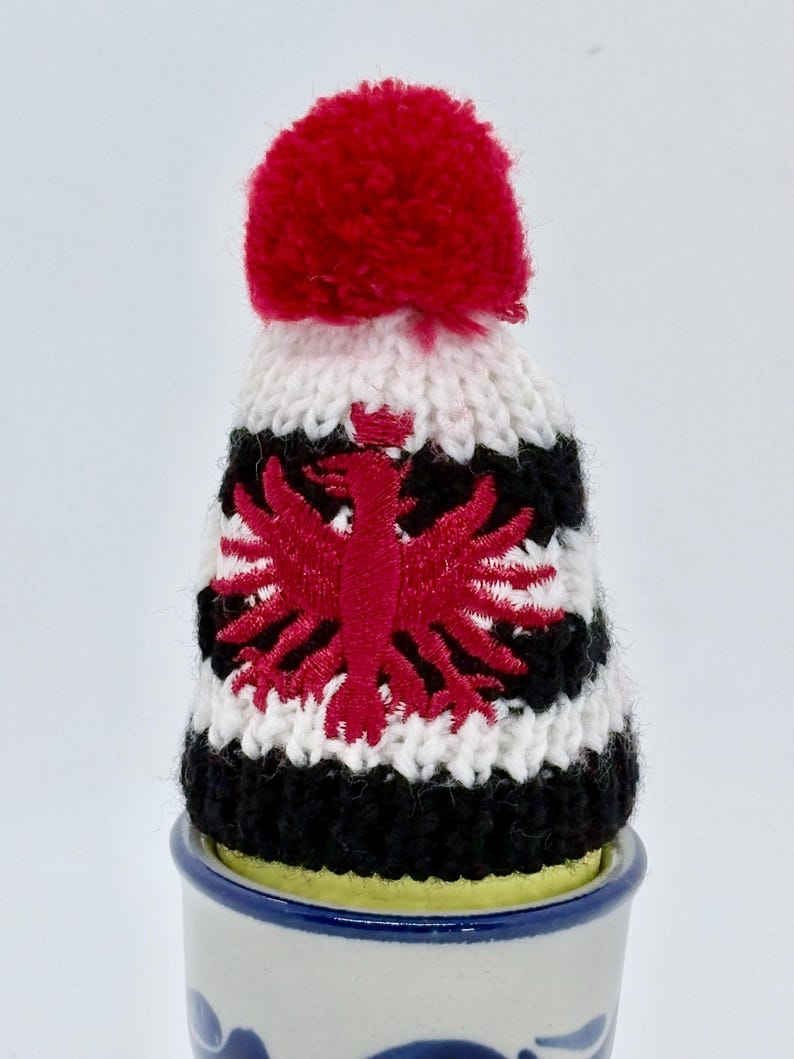 May include: A small knitted hat with a red pom-pom on top, featuring horizontal stripes of white and black yarn. The center of the hat displays a red embroidered eagle emblem. The hat is placed on top of a small, cylindrical ceramic container.